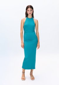 Robe midi turquoise à encolure halter dans un design ajusté. Tissu lisse, couleur unie sans motifs. Le modèle porte des sandales beiges à lanières.