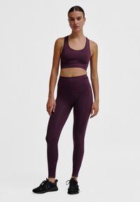 Fioletowy sportowy biustonosz i wysokowzwyżejce legginsy z elastycznej tkaniny, charakteryzujące się bezszwowym designem i subtelnymi akcentami logo. Czarne sneakersy dopełniają stylizację.