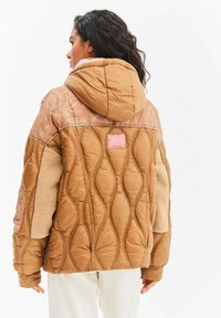 Kobieta o ciemnych kręconych włosach, ubrana w pikowaną brązową kurtkę z kapturem, z kwiatowymi wzorami na ramionach i beżowymi polarowymi łatami na rękawach.