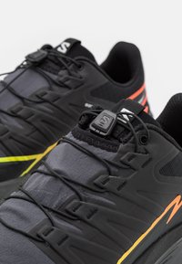 Scarpe da ginnastica nere con una tomaia testurizzata, design dinamico con accenti gialli e arancioni, e un sistema di lacci rapidi per una maggiore sicurezza.