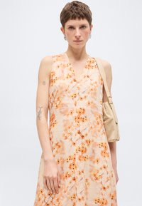 Γυναίκα με κοντά μαλλιά που φοράει αμάνικο κρεμ φόρεμα με πορτοκαλί λουλουδάτο σχέδιο και κρατάει μπεζ τσάντα ώμου.