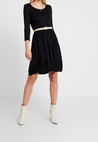 Robe noire côtelée avec une taille marquée, dotée d'une ceinture beige. Manches longues, jupe évasée et texture lisse. Portée avec des bottines blanches.