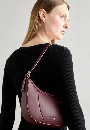 Borsa in pelle borgogna con una forma curva. Presenta hardware dorato e una superficie texturizzata, indossata sopra una maglietta nera a maniche lunghe con righe.