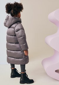 Geacă puffer de culoare gri deschis, cu glugă, având secțiuni matlasate. Botine de luptă negre cu șireturi și colanți gri completează ținuta.