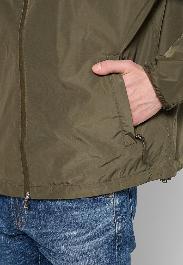 Olivegrüne, leichte Jacke mit glatter Textur, Reißverschluss vorne und Seitentaschen, getragen über blauen Jeans.