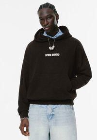 Sort hættetrøje med "STWD STUDIO" logo, frontlomme, båret over en blåstribet skjorte med krave, parret med lyse denim jeans.