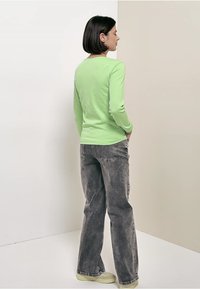 Vihreä, pitkähihainen puuvillapaita istuvalla leikkauksella, yhdistettynä väljälahkeisiin harmaisiin farkkuihin ja vaaleankeltaisiin tennareihin. Yksivärinen tausta.