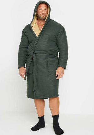 Homme portant un peignoir à capuche vert foncé avec ceinture et poches, debout pieds nus sur un fond blanc.