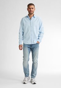 Světle modrá košile na knoflíky s dlouhými rukávy, v kombinaci s vybledlými modrými džíny a bílými teniskami. Košile má standardní límec a knoflíky.