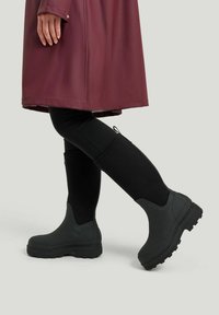 Mustat vedenkestävät nilkkurit, joissa on paksu pohja ja teksturoitu pinta, joita on käytetty mustien legginssien ja burgundin värisen sadetakin kanssa, seisoo harmaalla taustalla.