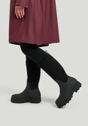 Mustat vedenkestävät nilkkurit, joissa on paksu pohja ja teksturoitu pinta, joita on käytetty mustien legginssien ja burgundin värisen sadetakin kanssa, seisoo harmaalla taustalla.
