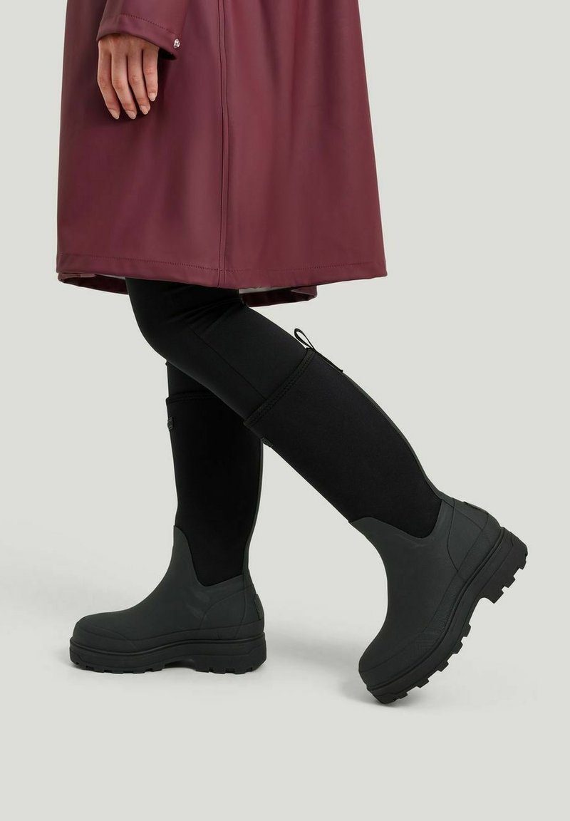 Mustat vedenkestävät nilkkurit, joissa on paksu pohja ja teksturoitu pinta, joita on käytetty mustien legginssien ja burgundin värisen sadetakin kanssa, seisoo harmaalla taustalla.