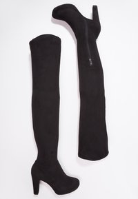 Botas negras de ante por encima de la rodilla con tacón alto. Cuentan con una cremallera lateral y una textura suave, extendiéndose hasta medio muslo.