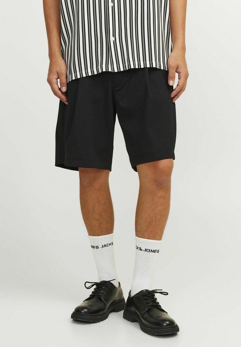 Muž v černých šortkách, černých šněrovacích botách, bílých ponožkách do poloviny lýtek s textem a tričku s černobílým pruhovaným vzorem, stojící na bílé podlaze.