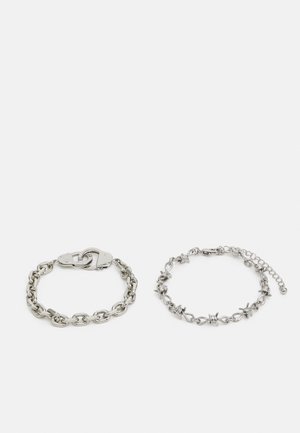 Pulseira de corrente prateada com fecho em forma de algemas à esquerda; à direita apresenta uma corrente decorativa com detalhes. Ambas têm um acabamento polido.