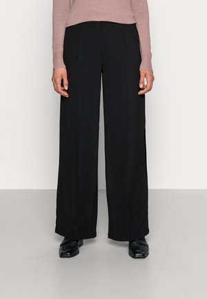 Schwarze, locker geschnittene Hosen mit Frontfalten aus einem glatten Stoff. Kombiniert mit einem figurbetonten, pinkfarbenen Langarmshirt und schwarzen Schuhen.