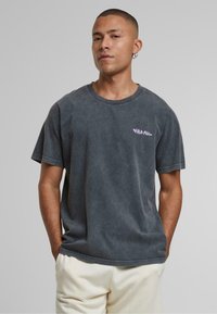 Sivý bavlnený tričko s uvoľneným strihom, krátkymi rukávmi, okrúhlym výstrihom a malou fialovou grafikou na hrudi. Hladká textúra.