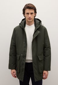 Giacca verde scuro con cappuccio, chiusura con zip e bottoni a scatto, due tasche frontali e un maglione bianco strutturato sotto. Tessuto liscio con finitura opaca.