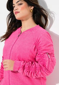Chaqueta rosa brillante con cremallera, con detalle fruncido en las mangas y un bolsillo lateral. Hecha de una tela suave con un acabado texturizado.
