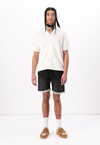 Short-sleeve white button-up shirt paired with black denim shorts, rolled cuffs, white socks, and brown sneakers, worn by a man.