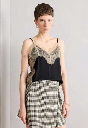 Femme aux cheveux courts portant des boucles d'oreilles créoles dorées, un caraco noir avec une dentelle beige et une jupe à taille haute à carreaux avec des boutons latéraux, debout.