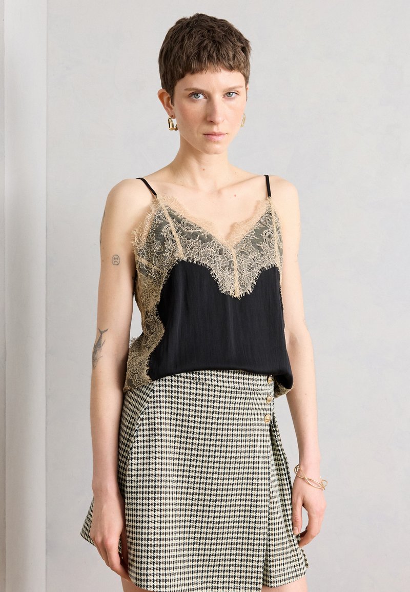 Γυναίκα με κοντά μαλλιά που φορά χρυσούς κρίκους σκουλαρίκια, μαύρο τοπ με μπεζ δαντέλα και καρό ψηλόμεση φούστα με πλαϊνά κουμπιά, όρθια.