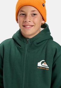 Sudadera verde con cremallera y textura suave, con un logotipo blanco y detalles en naranja. Combinada con un gorro naranja de canalé.