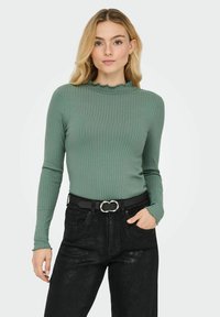 Groene geribbelde longsleeve met een geschulpte halslijn, gecombineerd met zwarte high-waisted jeans en een zwarte riem met een zilveren gesp in de vorm van een schakeltje.