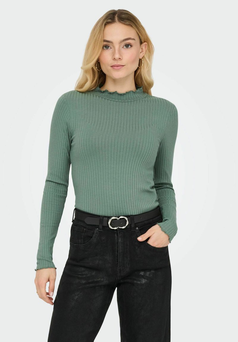 Groene geribbelde longsleeve met een geschulpte halslijn, gecombineerd met zwarte high-waisted jeans en een zwarte riem met een zilveren gesp in de vorm van een schakeltje.