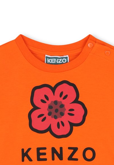 Oranje katoenen T-shirt met een zwarte omtrek van een roze bloem met donkerroze accenten en knoopdetails op de schouder. Merken zichtbaar eronder.
