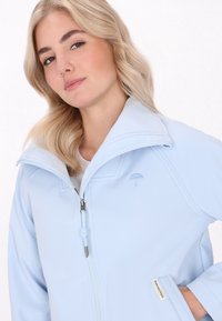 Donna bionda sorride, indossando una giacca azzurro chiaro con un piccolo logo a forma di ombrello. Lo sfondo bianco mette in risalto i suoi tratti e il suo abbigliamento.