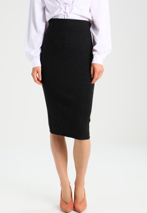 Vrouw met een witte blouse, een zwarte kokerrok tot op de knie en koraalkleurige puntige pumps, staand voor een effen achtergrond.