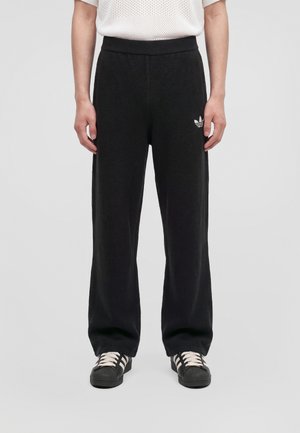 Le mannequin porte un pantalon de survêtement Adidas noir à jambes larges, avec une taille élastique et le logo Trefoil blanc, associé à des baskets noires à rayures.