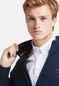 Jūrinė zip-up striukė su įtemtų rankogalių, oranžinės spalvos akcentas, dėvima ant baltos marškinėlių su apykakle. Detalus modelio, laikant striukės užtrauktuką, vaizdas.