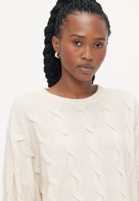 Krema pulover s kabelskim pletenjem, s okruglim izrezom i opuštenim krojem, prikazuje podignute teksturirane uzorke. Glatka tkanina i ležeran dizajn.