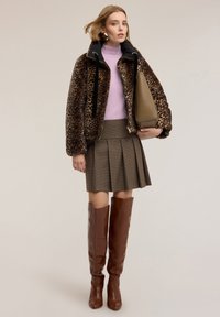 Kobieta ubrana w kurtkę w panterkę, lawendowy sweter, plisowaną spódnicę w kratę, trzyma beżową torbę i nosi brązowe skórzane kozaki za kolano.