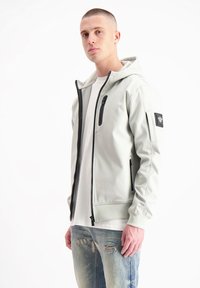 Lichtgrijze capuchonjack gemaakt van zacht materiaal, met een frontale rits, zijzakken en een zwarte ritsaccent. Draagt over een wit shirt.