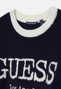 Pull en tricot bleu marine avec col côtelé crème. Présente un logo "GUESS" blanc audacieux et le texte "Los Angeles". Texture douce, coupe décontractée.