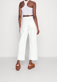 Kvinna som står vänd bort med vita vida cropped byxor, vit racerback crop top och bruna plattformsskor utan snörning.