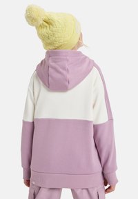 Sudadera con bloque de color púrpura y crema, con capucha y puños de canalé. Presenta un tejido suave y un pequeño detalle de logotipo en el dobladillo.