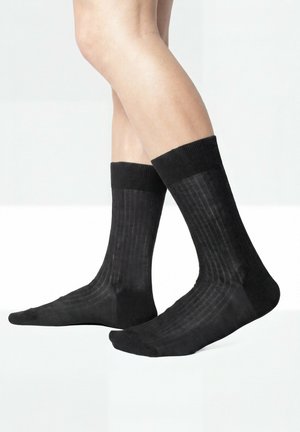 Beine, die schwarze gerippte Socken bis zur Mitte der Wade mit Bündchen tragen, stehend auf einer weißen Oberfläche vor einem hellgrauen Hintergrund.