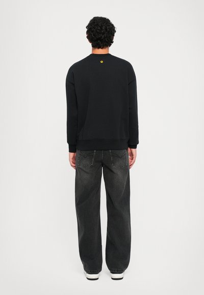 Felpa nera con scollo rotondo e logo giallo sul retro, abbinata a jeans grigi scuri a vestibilità morbida e sneakers bianche.