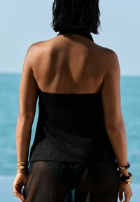 Černý halter top s texturou a přiléhavým korzetem, kombinovaný s průhlednými černými kalhotami. Šperky zahrnují několik náramků na zápěstí.