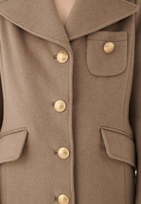 Manteau en polaire marron avec un grand col, des boutons dorés et une poche à rabat. Il présente un tissu texturé et un design structuré.