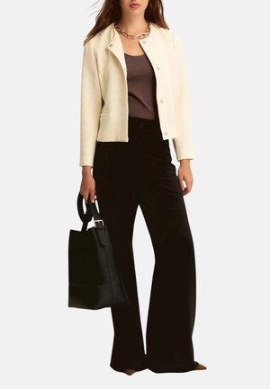 Donna che indossa una giacca crema texturizzata, top marrone, pantaloni neri a gamba larga, scarpe marroni, collana a catena gioiello, che tiene una borsa nera.