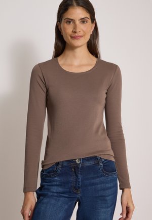 Femme aux longs cheveux foncés portant un haut marron ajusté à manches longues et un jean bleu, debout devant un fond uni.
