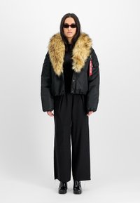 Svart cropped jakke med fuskepelskrage. Jakken har en glatt tekstur og store ermer. Modellen har på seg svarte vide bukser og solbriller.