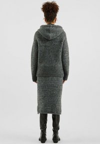 Woman with curly hair wearing a grey knit hooded sweater and matching long skirt, paired with black knee-high boots, standing facing away.