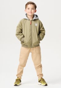 Olivgrüne Bomberjacke mit grauem Kapuzenumschlag, kombiniert mit hellbeigen Jogginghosen und gemusterten Sneakers in Gelb, Braun und Weiß.