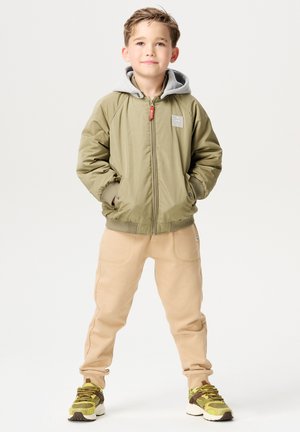 Olijfgroen bomberjack met een grijze capuchon, gecombineerd met lichtbeige sweatpants en gestructureerde sneakers in geel, bruin en wit.
